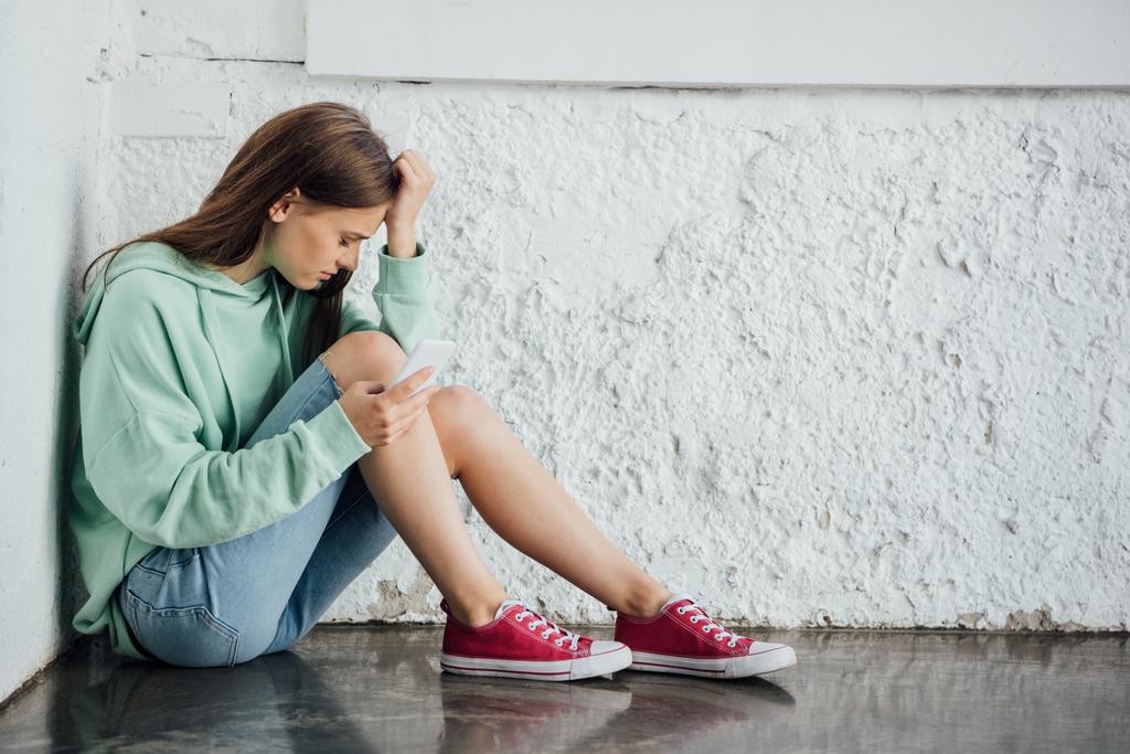smutná dívka, která sedí u stěny s texturou a drží telefon Smartphone - Fotografie, Obrázek