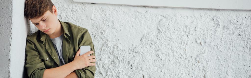 πανοραμικό σφηνάκι λυπημένο έφηβο κρατώντας smartphone κοντά σε υφή τοίχου - Φωτογραφία, εικόνα