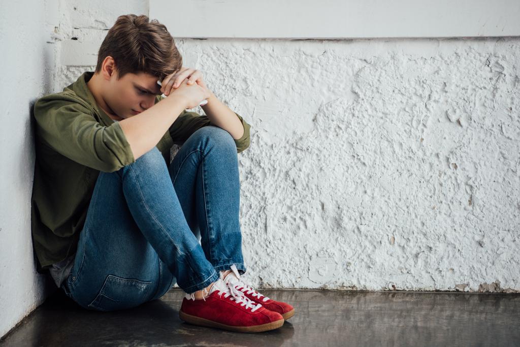 сумний підліток в джинсах тримає смартфон і сидить на підлозі
 - Фото, зображення
