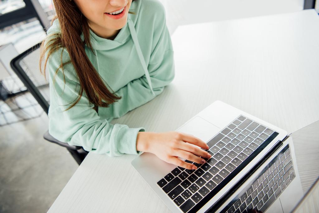 vista parcial de la chica sonriente en sudadera con capucha sentado en la mesa y escribiendo en el teclado del ordenador portátil
 - Foto, imagen