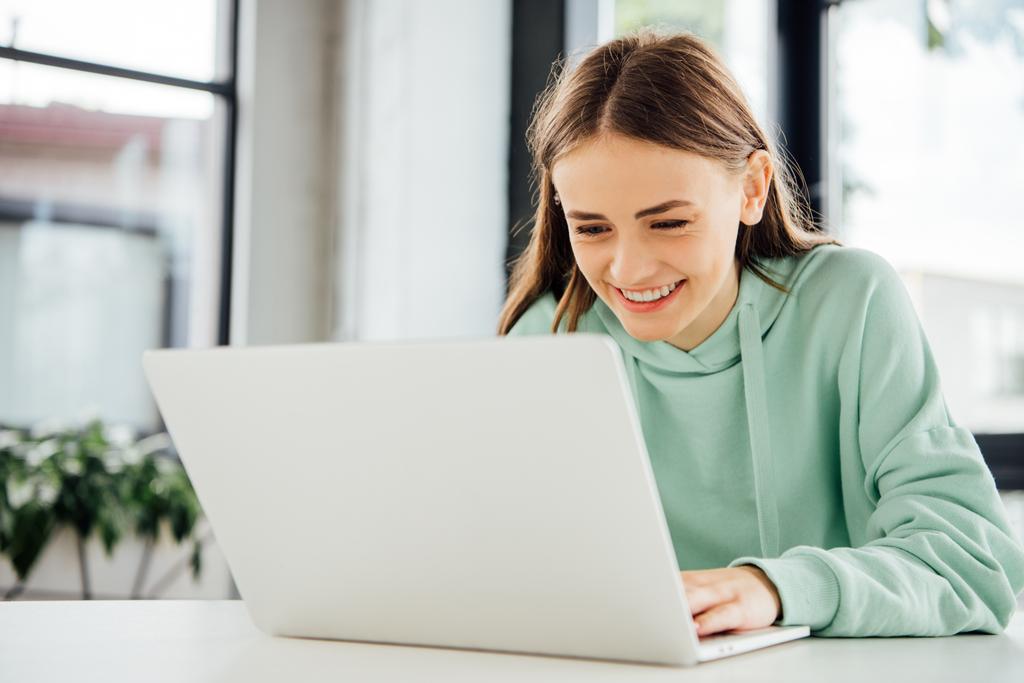 χαμογελαστός κορίτσι σε casual φούτερ με κουκούλα κάθεται στο τραπέζι και χρησιμοποιώντας το laptop - Φωτογραφία, εικόνα