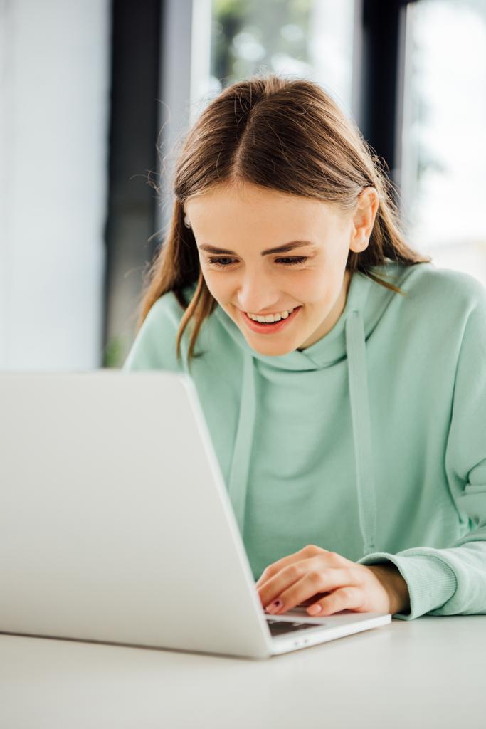gündelik kapüşonlu gülümseyen kız masada oturan ve dizüstü bilgisayar kullanarak - Fotoğraf, Görsel