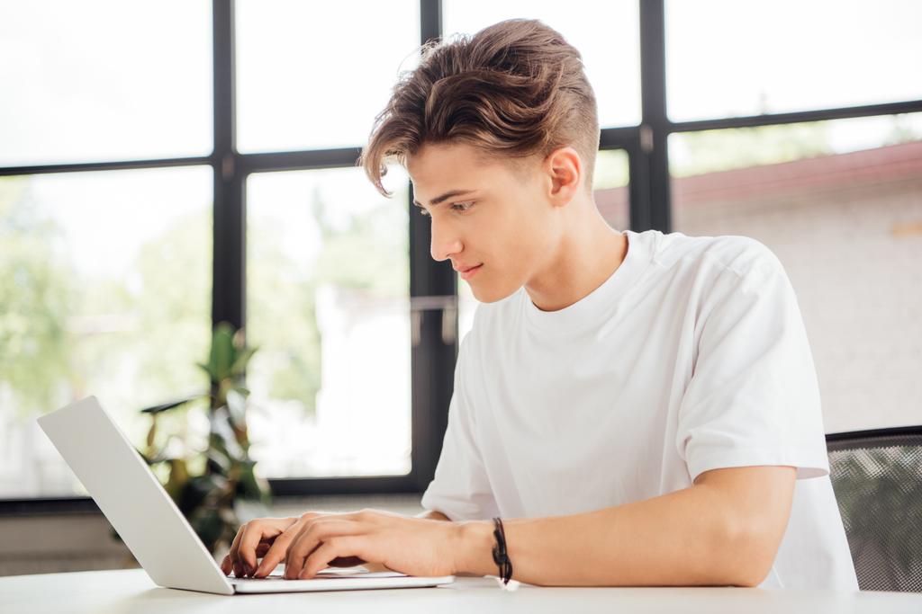 enfocado adolescente chico en blanco camiseta usando portátil en casa
 - Foto, imagen