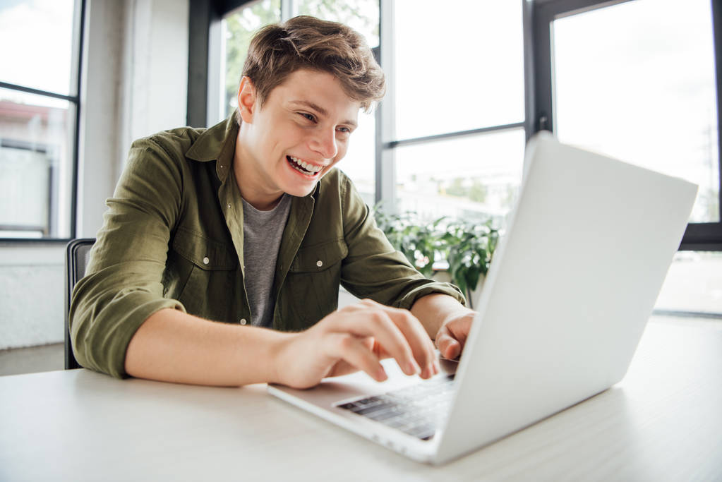 opgewonden tiener jongen zitten aan tafel en typen op laptop toetsenbord thuis - Foto, afbeelding
