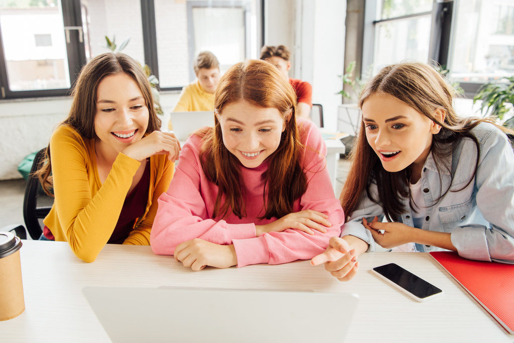 drie glimlachende meisjes kijken naar laptop scherm op school - Foto, afbeelding