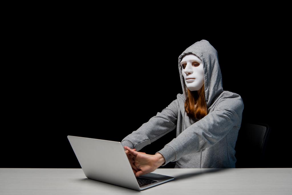 ανώνυμο κορίτσι με μάσκα και κουκούλα καθισμένος κοντά στο φορητό υπολογιστή και τεντώνοντας τα χέρια κατά τη διάρκεια του κυβερνοεκφοβισμού απομονωθεί σε μαύρο - Φωτογραφία, εικόνα