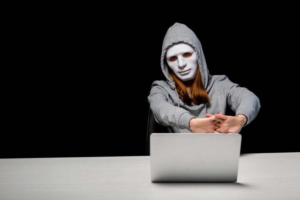 анонімна дівчина в масці і светрі сидить біля ноутбука і розтягує руки під час кіберзалякування ізольовано на чорному
 - Фото, зображення