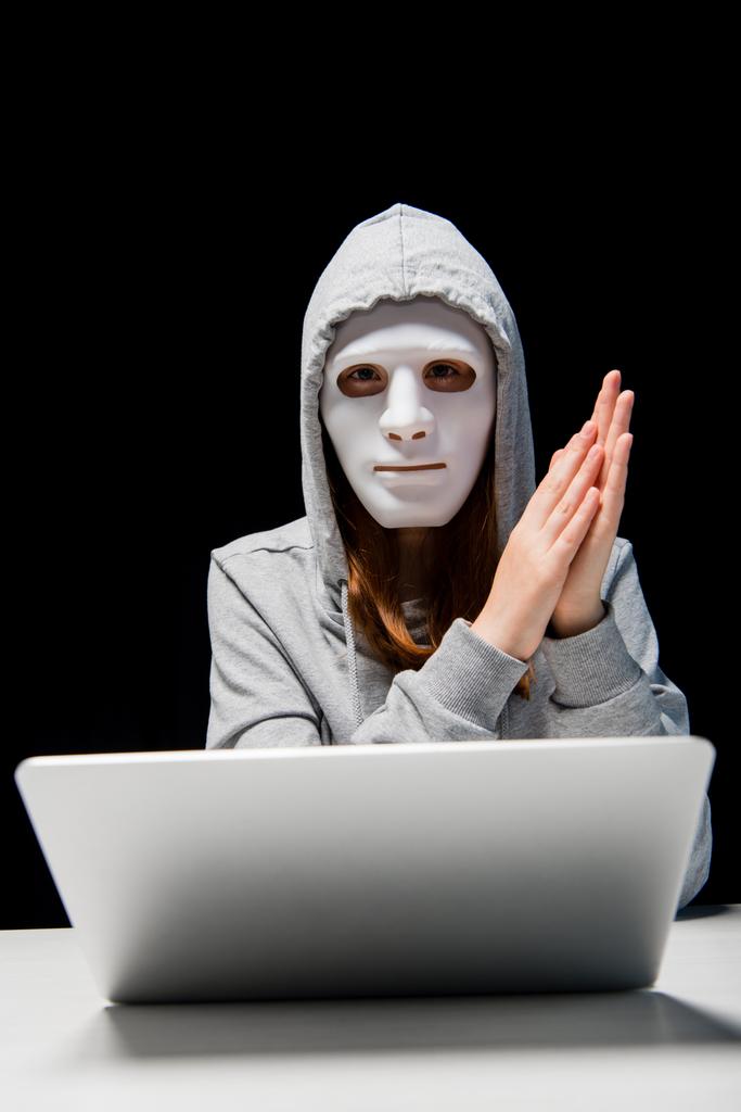 vista frontal de una chica anónima con máscara y sudadera con capucha sentada cerca de la computadora portátil y frotándose las manos durante el cyberbullying aislado en negro
 - Foto, Imagen