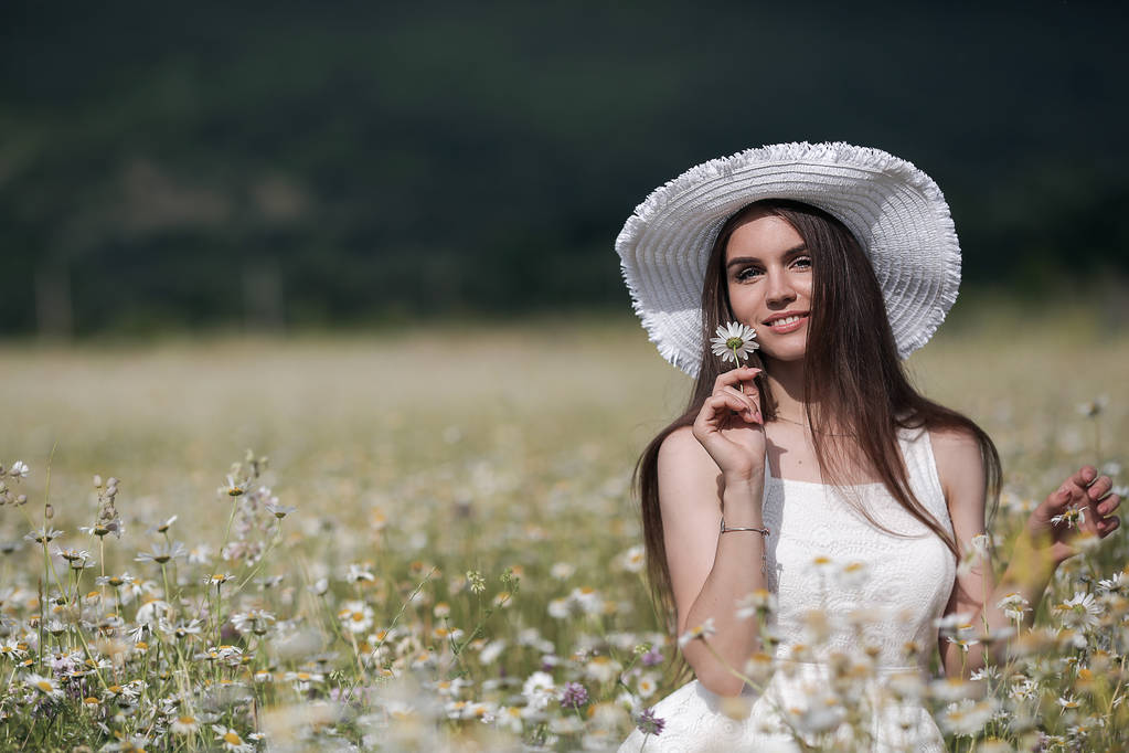 自然を楽しむ白いヒナギクのフィールドに花の花束を持つ屋外の美しい女の子。花、太陽の光と夏のフィールドに楽しみを持っている白いドレスで長い髪を持つ美しいモデル。春の草原、田舎の若い幸せな女性 - 写真・画像