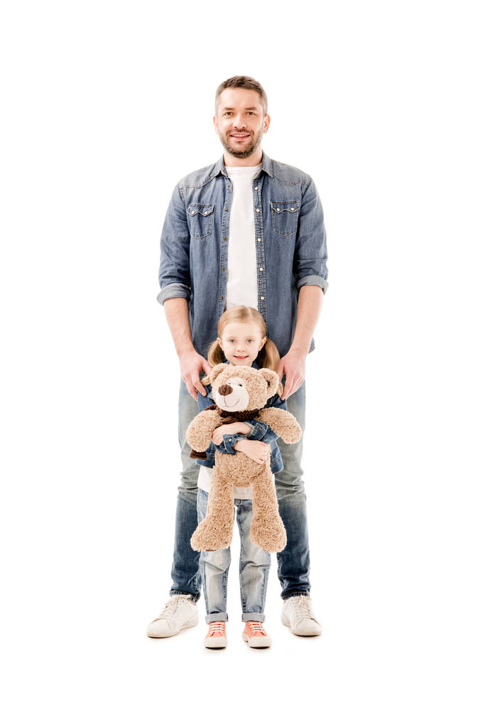 Вид в полный рост улыбающегося папы в джинсах и дочери с плюшевым медведем, изолированным на белом
 - Фото, изображение
