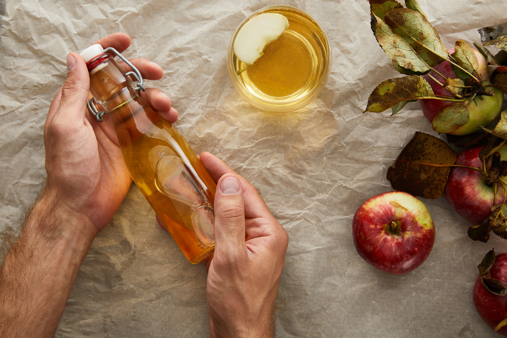 περικομμένη εικόνα του ανθρώπου που κρατά ένα μπουκάλι μηλίτη μήλου κάτω από λαδόκολλα με μήλο και γυαλί - Φωτογραφία, εικόνα