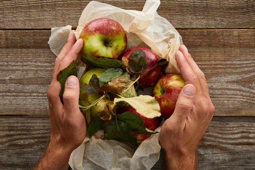 περικομμένη εικόνα του ανθρώπου που κρατά λαδόκολλα με μήλα σε ξύλινη επιφάνεια - Φωτογραφία, εικόνα