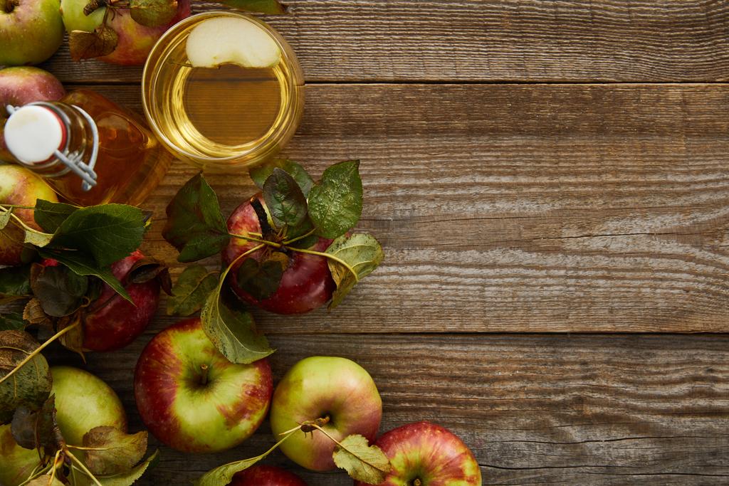 Draufsicht auf reife Äpfel in Flaschennähe und Glas frischer Apfelwein auf Holzoberfläche - Foto, Bild