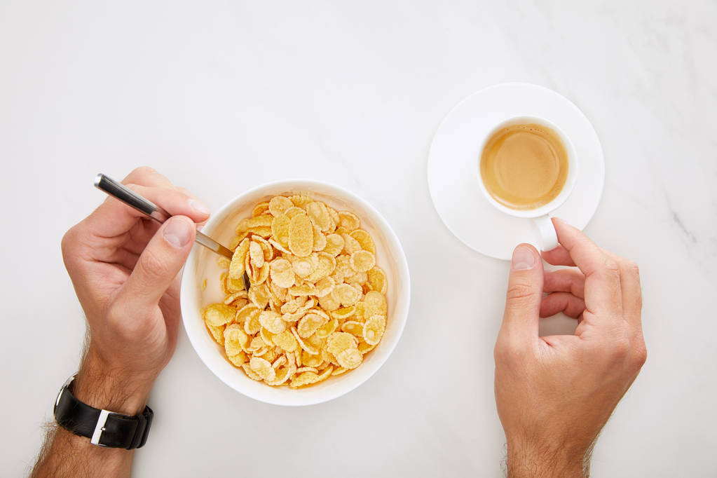 abgeschnittenes Bild des Mannes mit Tasse Kaffee und Löffel in Schüssel mit Cornflakes auf weißem Marmorhintergrund - Foto, Bild