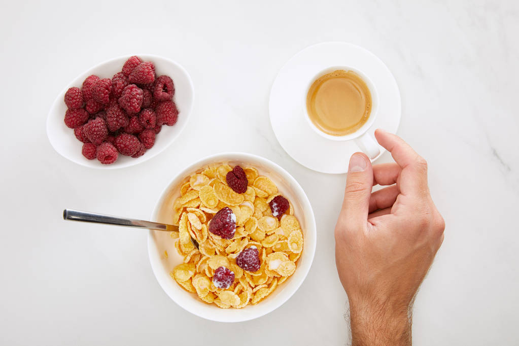cropped image of man holding cup of coffee near bowl with cornflakes and plate with fresh raspberry on white marble background - Photo, Image