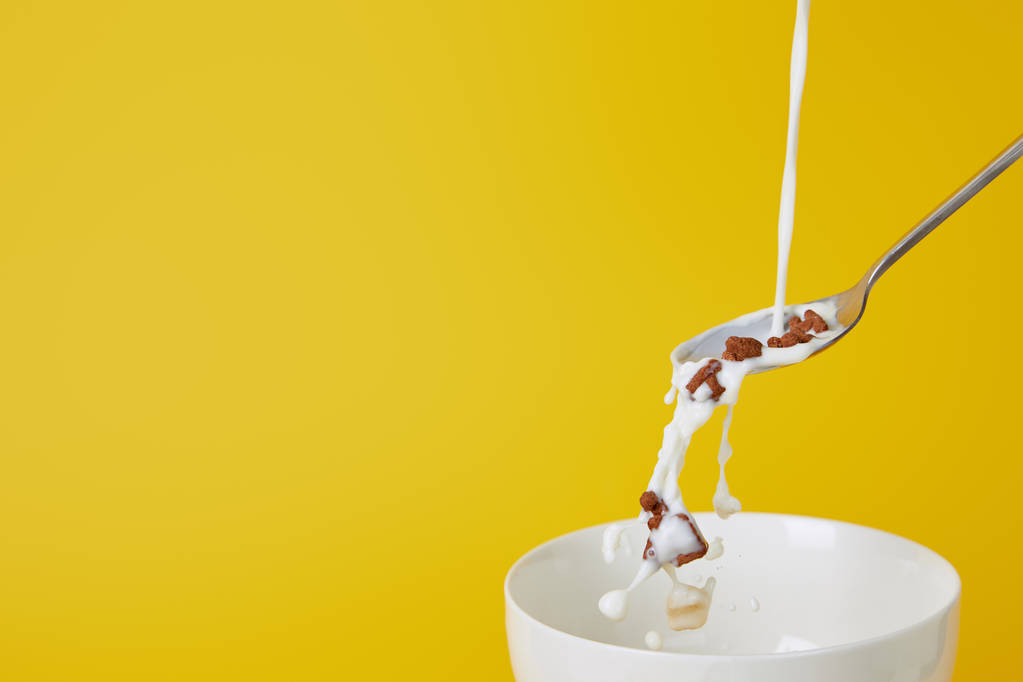 łyżeczka z wyborem płatków zbożowych i rozpryskami mleka powyżej pustej miski na żółto - Zdjęcie, obraz