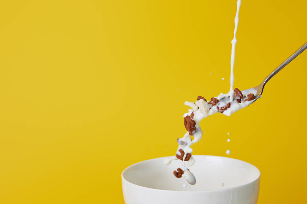 ложка с разнообразными хлопьями и брызги молока над миской изолированы на желтый
 - Фото, изображение