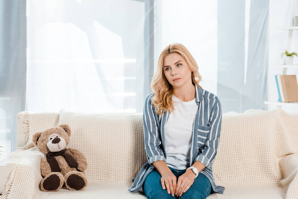 upset woman sitting on sofa near soft toy at home  - Photo, Image