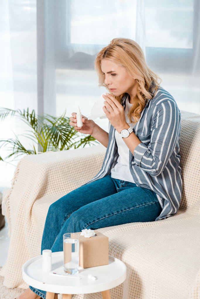 Kranke Frau sitzt auf Sofa und hält Nasenspray und Gewebe in der Hand  - Foto, Bild