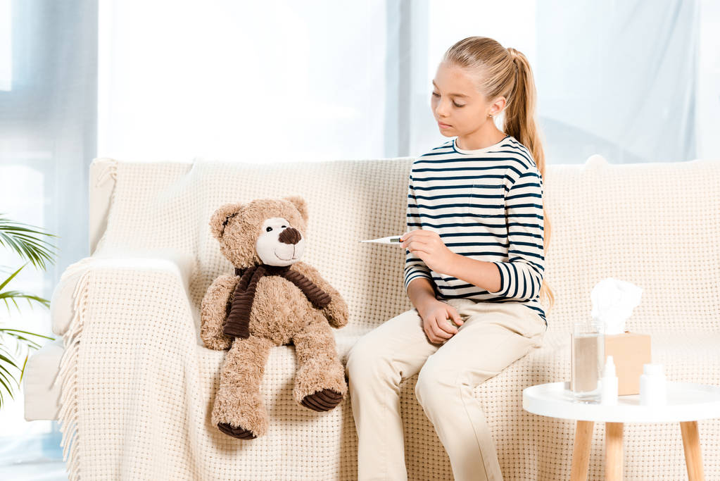 dítě držící digitální teploměr poblíž medvídka a sedícího na pohovce  - Fotografie, Obrázek