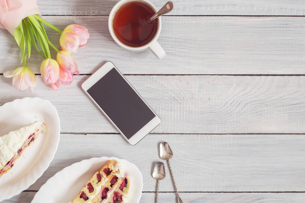 Ciasto, telefon, filiżanka herbaty i różowe tulipany na białym drewnianym stole - Zdjęcie, obraz