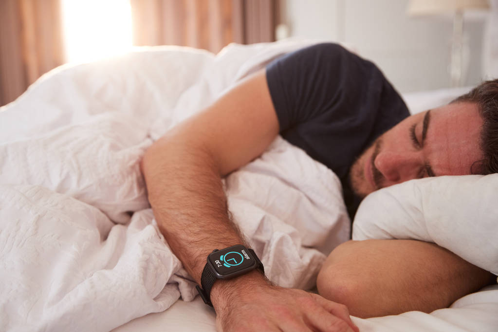 Άνθρωπος κοιμάται στο κρεβάτι φορώντας έξυπνο ρολόι καθώς ο ήλιος σπάει μέσα από κουρτίνες - Φωτογραφία, εικόνα