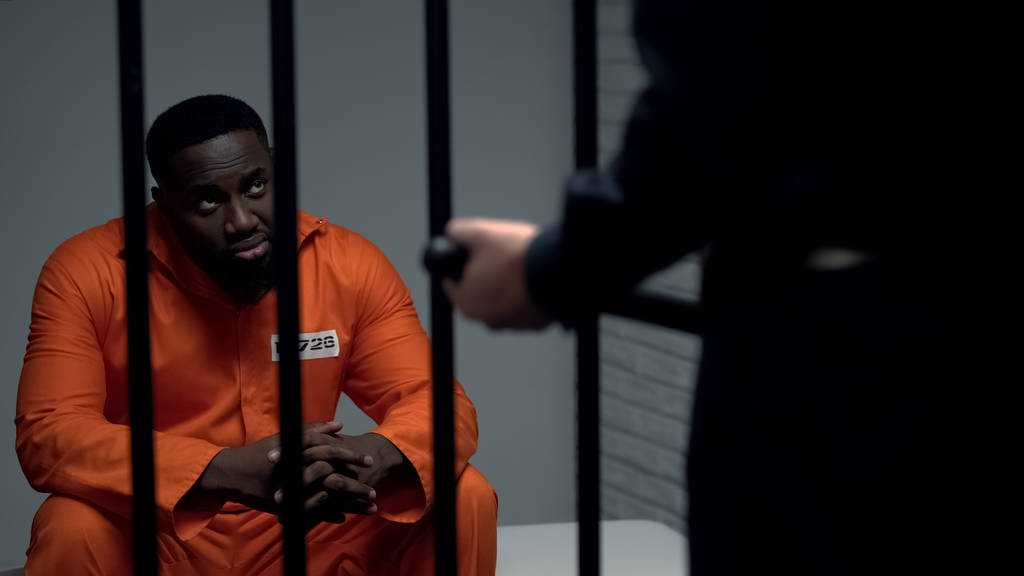 Gefängniswärter mit Schlagstock sieht afroamerikanischen Häftling in Zelle an, Belästigung - Foto, Bild