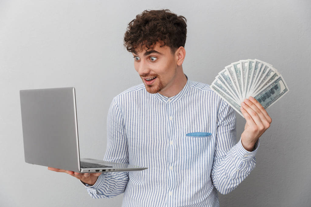 Porträt eines entzückten kaukasischen Mannes im Hemd, der in die Kamera lächelt, während er einen silbernen Laptop und einen Haufen Bargeld in der Hand hält - Foto, Bild