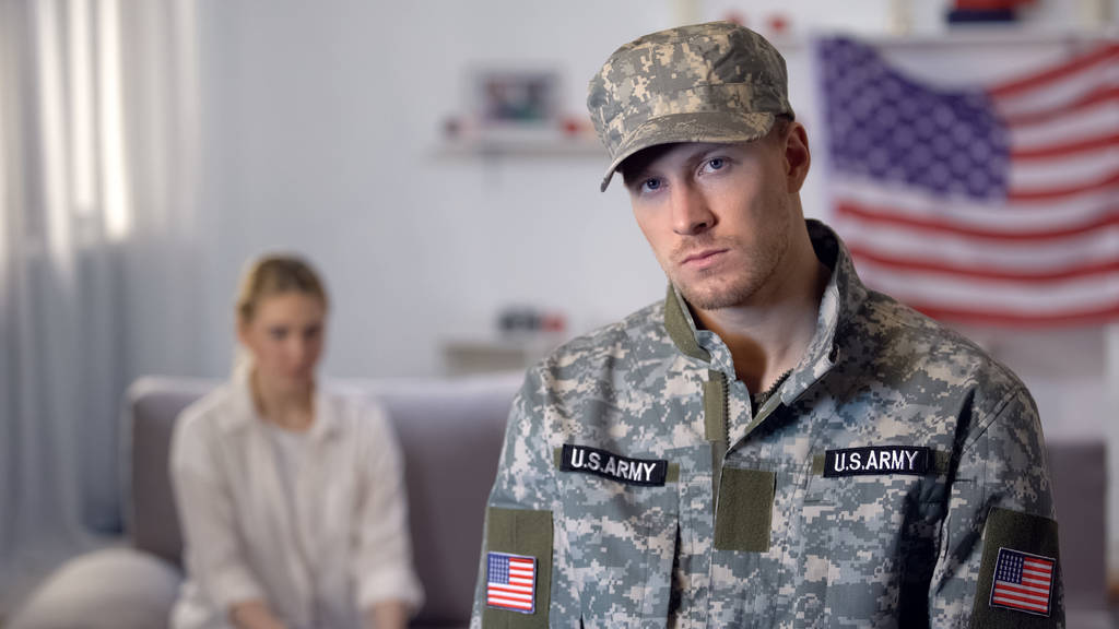 Loukkaantunut amerikkalainen sotilas katselee kameraa, järkyttynyt vaimo istuu sohvalla, riitaa
 - Valokuva, kuva