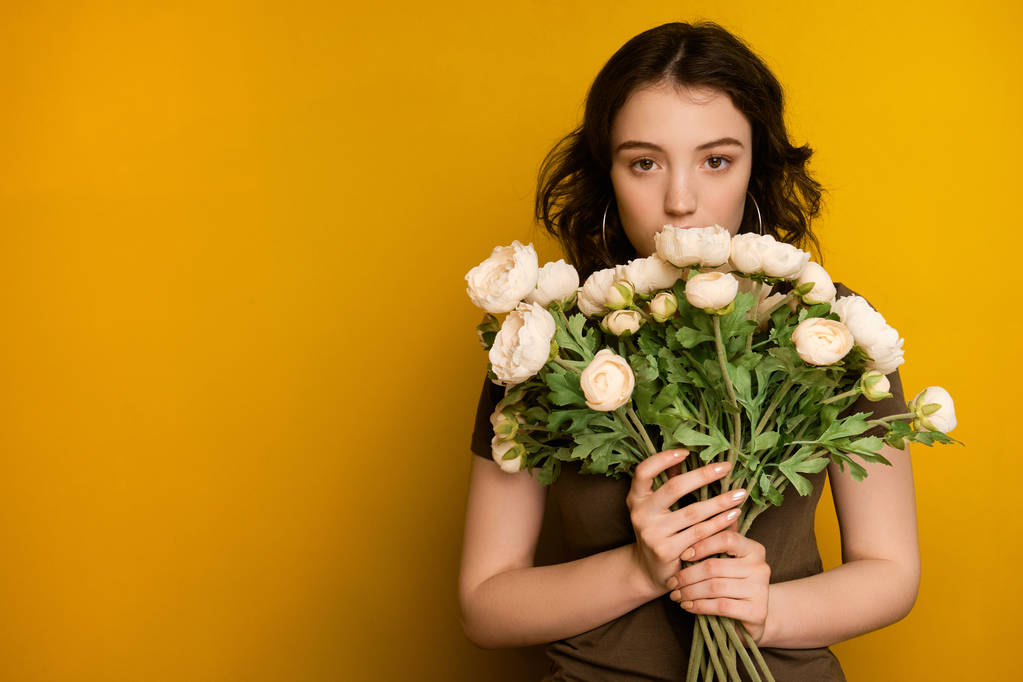 Une fille aux cheveux bruns bouclés se tient sur un fond jaune et regarde la caméra par derrière un bouquet de fleurs
. - Photo, image