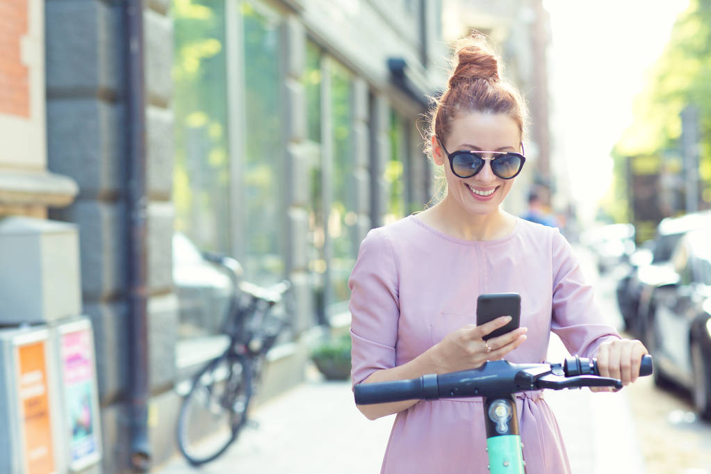 Szczęśliwa Kobieta w okulary przeciwsłoneczne za pomocą smartfona i wynajęcie nowoczesnego skuter elektryczny z aplikacją - Zdjęcie, obraz