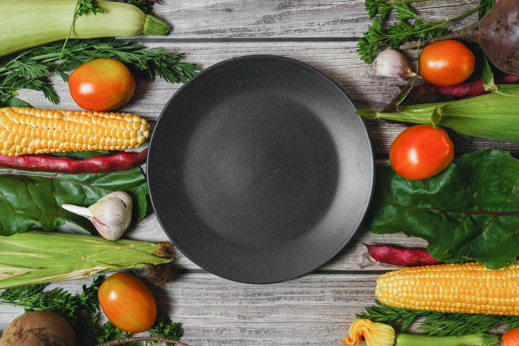 Creative layout made of corn, tomatoes, zucchini, carrot, beetroot, garlic, bean and black plate on wooden background. Food concept. Flat lay, top view. Free space for your text. - Photo, Image