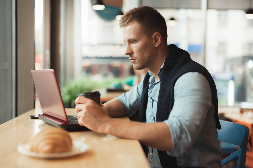 junger schöner Mann arbeitet in seinem Laptop im Café, isst Croissant und trinkt warmen Kaffee zum Mittagessen - Foto, Bild