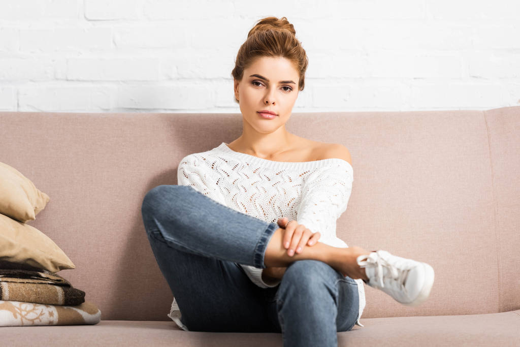 ελκυστική γυναίκα σε λευκό πουλόβερ κοιτάζοντας την κάμερα και κάθεται στον καναπέ  - Φωτογραφία, εικόνα