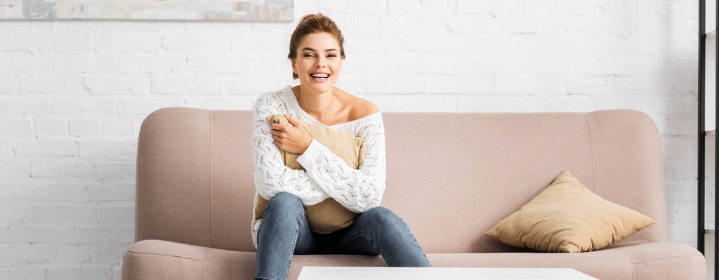 panoramic shot of attractive woman in white sweater holding pillow and looking at camera  - Photo, Image