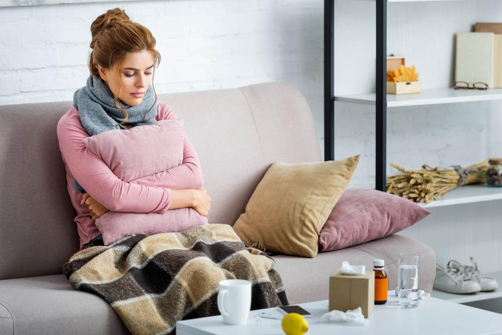 femme attrayante et malade avec écharpe grise assise sur le canapé et tenant oreiller
  - Photo, image
