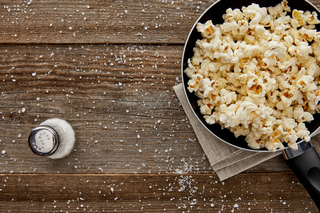 Draufsicht auf frisches Popcorn in Pfanne mit Salz auf Holzgrund - Foto, Bild
