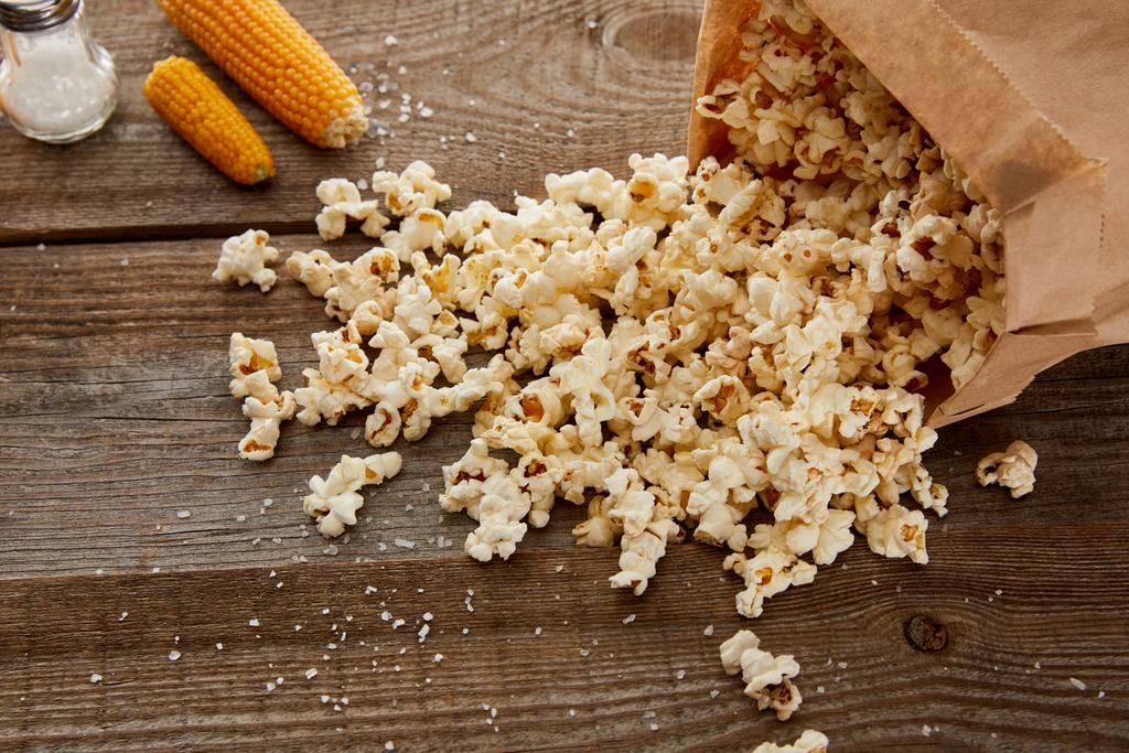 Соленый вкусный попкорн разбросан из бумажного пакета рядом с кукурузой на деревянном фоне
 - Фото, изображение