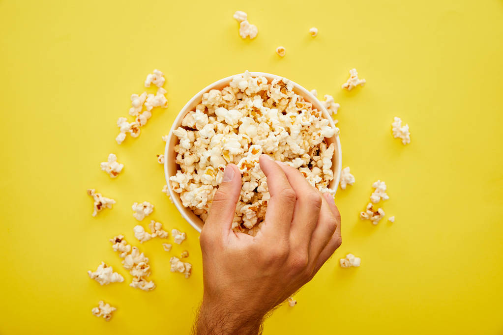gedeeltelijke weergave van de mens het nemen van heerlijke verse popcorn uit emmer op gele achtergrond - Foto, afbeelding