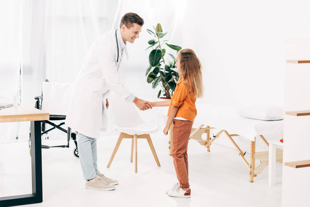 visão de comprimento total do pediatra em casaco branco e crianças apertando as mãos e olhando um para o outro
 - Foto, Imagem