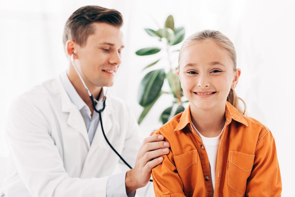 Lächelnder Kinderarzt im weißen Kittel untersucht Kind mit Stethoskop - Foto, Bild