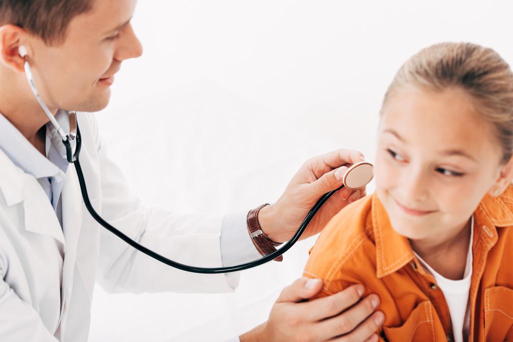 pediatrist σε λευκό παλτό εξέταση παιδί με στηθοσκόπιο απομονώθηκε σε λευκό - Φωτογραφία, εικόνα
