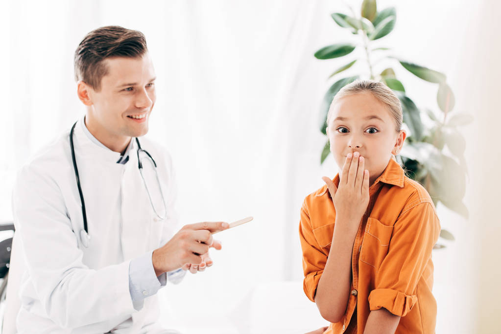 παιδί καλύπτοντας το στόμα με το χέρι και χαμογελαστή pediatrist κρατώντας ιατρική σπάτουλα στην κλινική - Φωτογραφία, εικόνα
