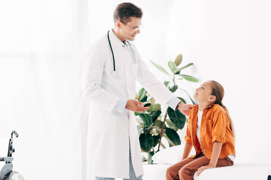 улыбающийся педиатр в белом халате и ребенок смотрят друг на друга в клинике
 - Фото, изображение