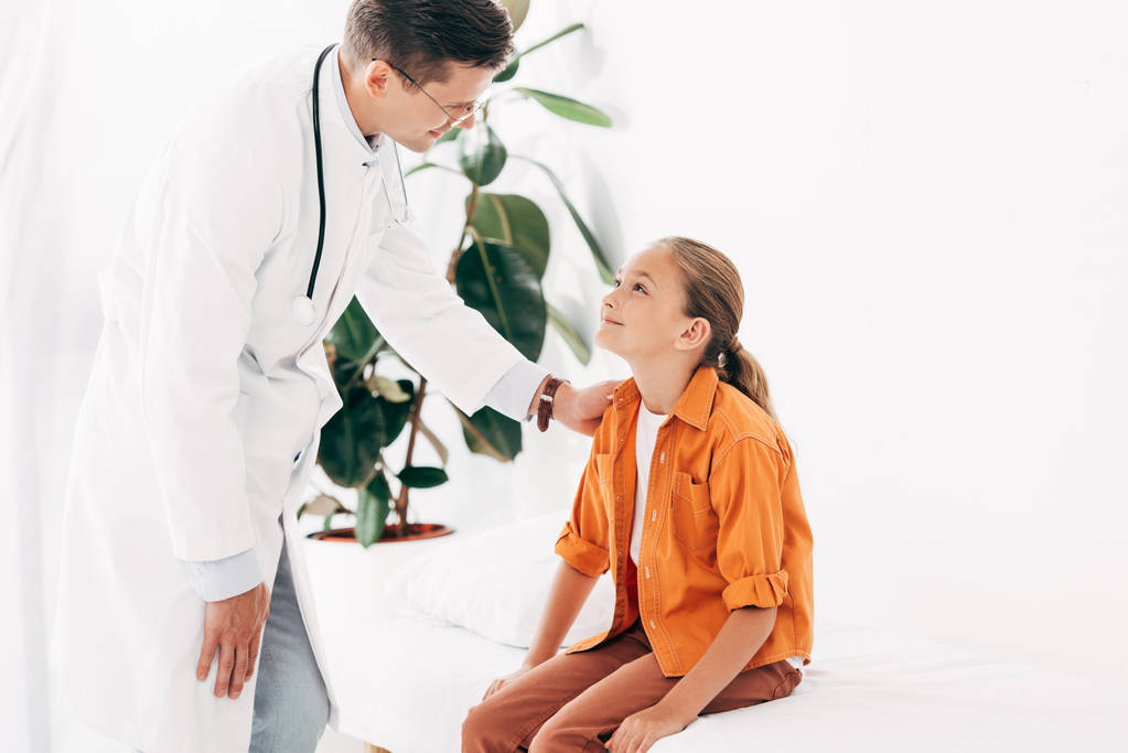 smiling pediatrist in white coat and child looking at each other in clinic - Photo, Image