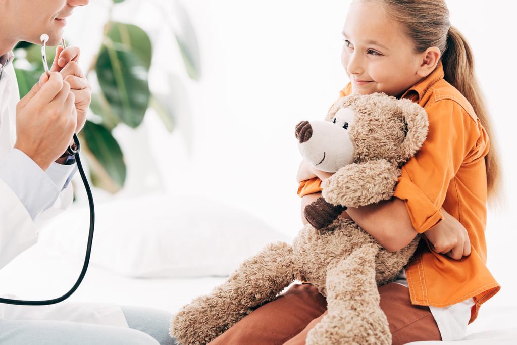 Teilansicht von Kinderarzt in weißem Mantel und Kind mit Teddybär in Klinik - Foto, Bild