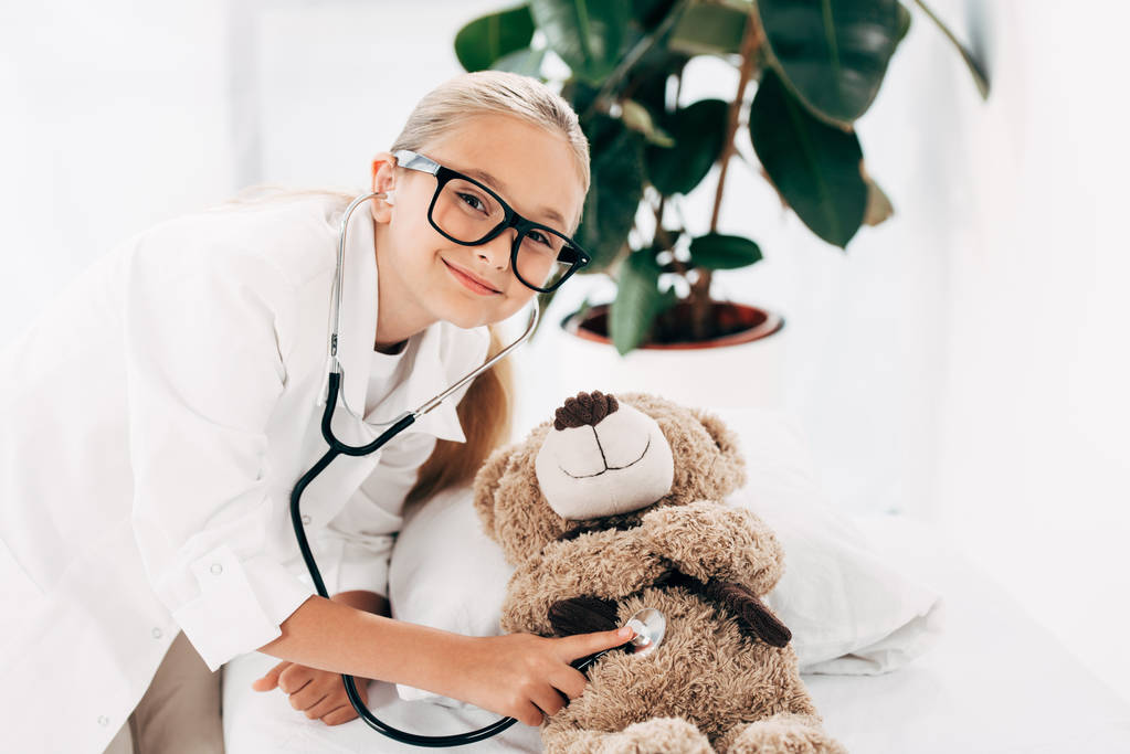Lächelndes Kind im Arztkostüm untersucht Teddybär mit Stethoskop - Foto, Bild