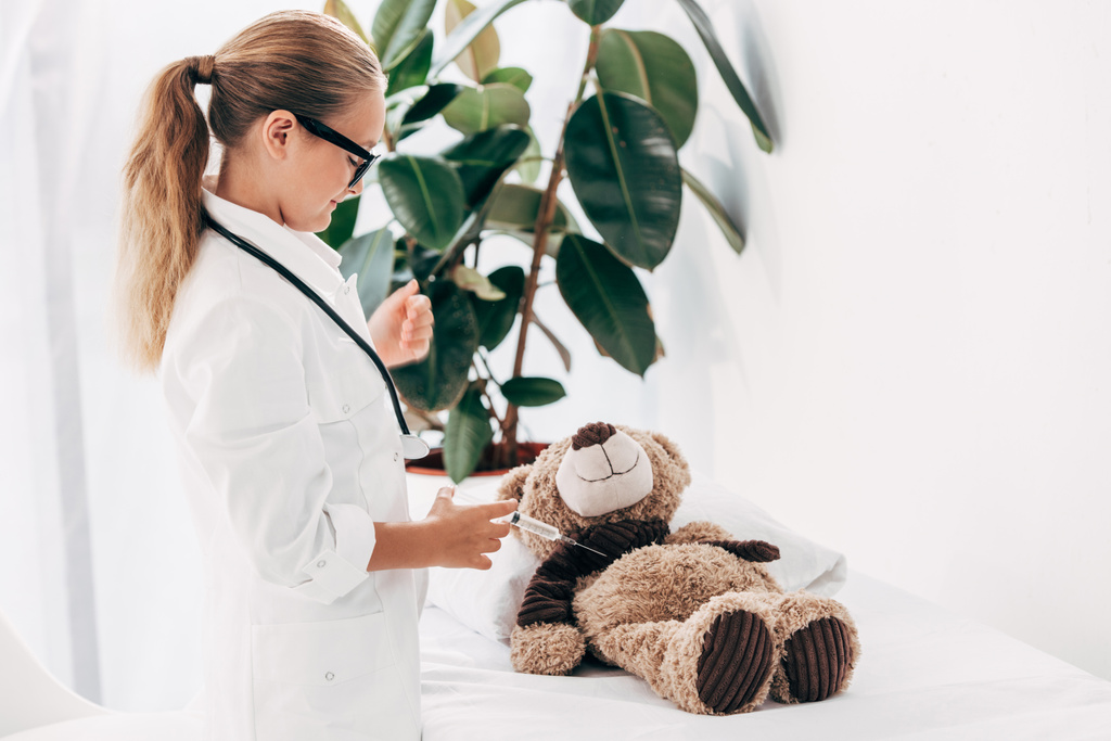 kind in Doctor kostuum en glazen staande in de buurt van Teddy Bear en het houden van de spuit - Foto, afbeelding