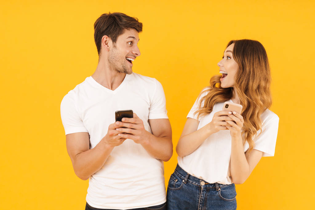 Πορτρέτο του όμορφο ζευγάρι άντρας και γυναίκα σε βασικά μπλουζάκια κοιτάζοντας ο ένας τον άλλο, κρατώντας smartphone - Φωτογραφία, εικόνα