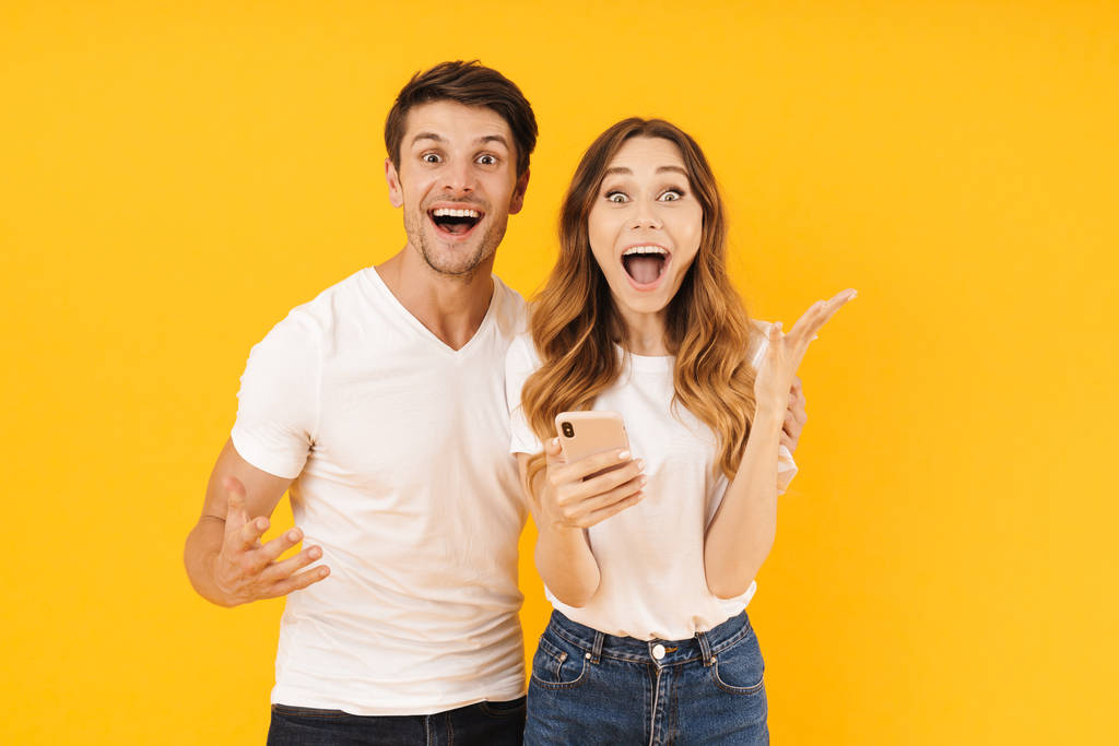 Portrait de couple excité homme et femme en t-shirts de base se réjouissant tout en se tenant debout avec smartphone
 - Photo, image